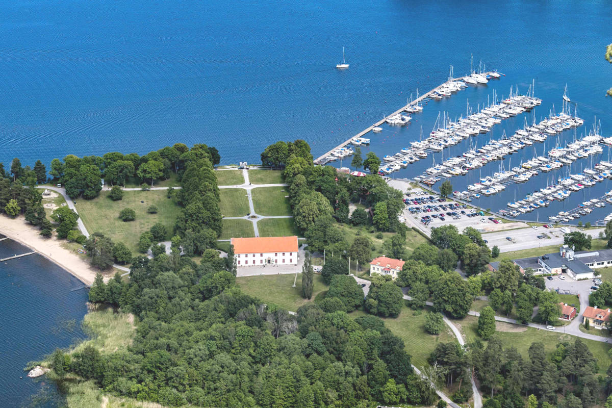 Veduta aerea di un porto turistico con molte barche a vela attraccate vicino al castello di Sundbyholm, adiacente a un parco alberato, edifici e una spiaggia accanto a uno specchio d'acqua blu.