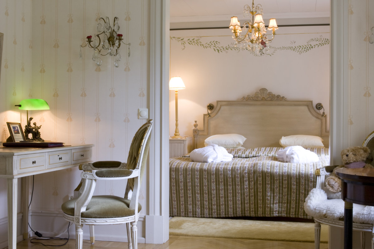 La camera da letto in stile vintage del Castello di Sundbyholm dispone di un letto matrimoniale con testiera decorata, due accappatoi, lampade da comodino, una scrivania con sedia e lampada verde e un lampadario appeso al soffitto.