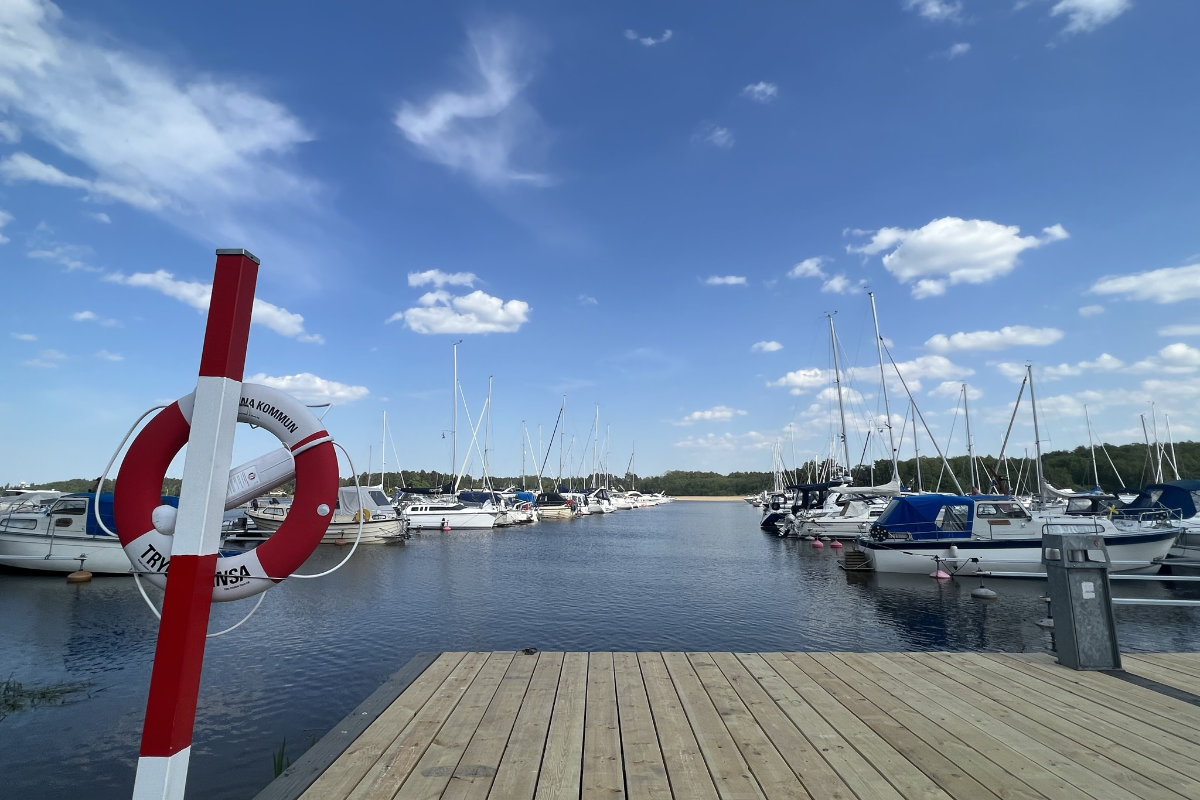 Un molo di legno si affaccia su un porto turistico vicino al castello di Sundbyholm, con numerose barche a vela e a motore ormeggiate sotto un cielo azzurro con nuvole sparse. Un salvagente è montato su un palo a sinistra.