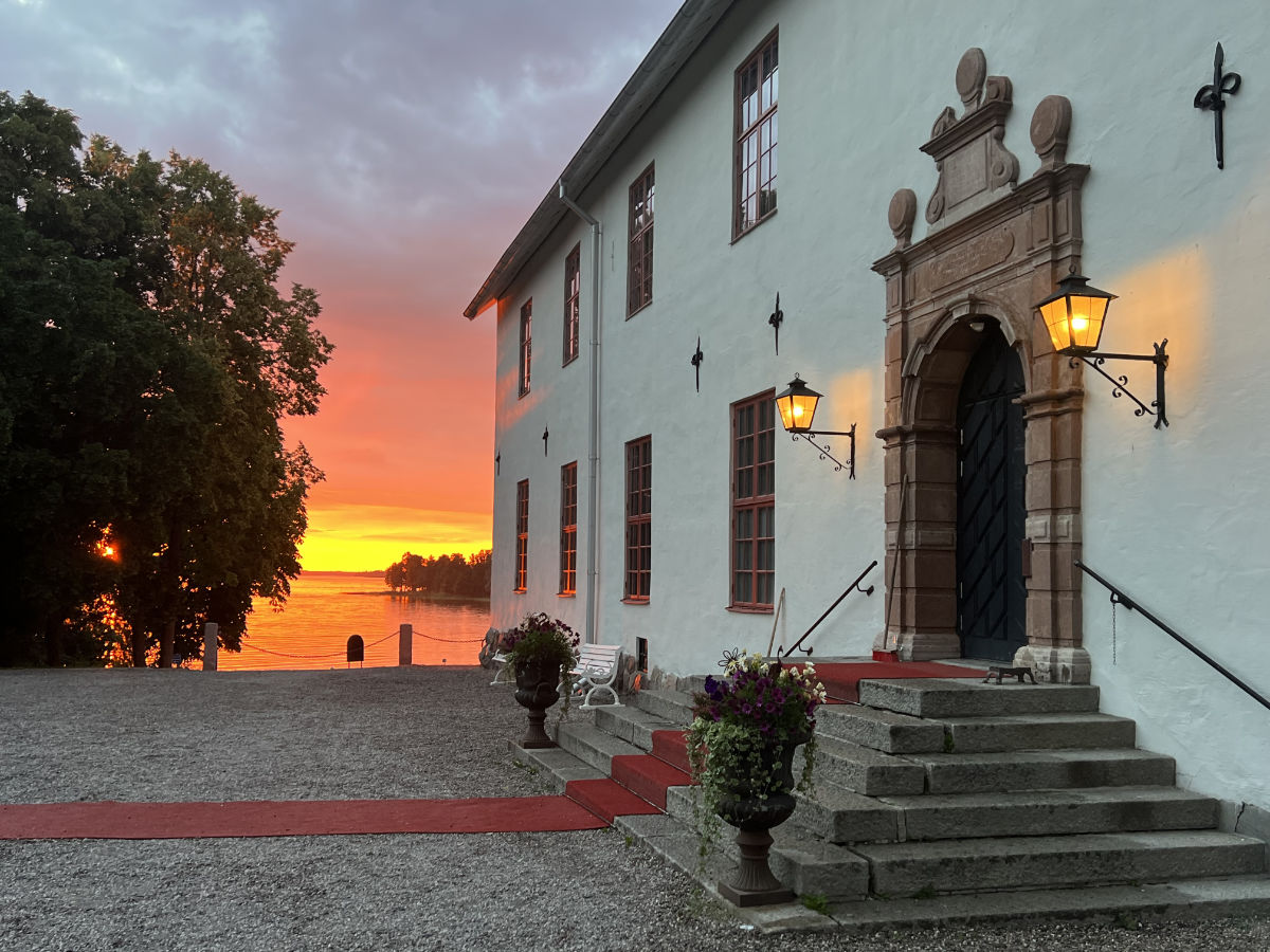 L'edificio storico del Castello di Sundbyholm è caratterizzato da gradini in pietra, un tappeto rosso e lanterne luminose al tramonto, con acqua e alberi visibili sullo sfondo.