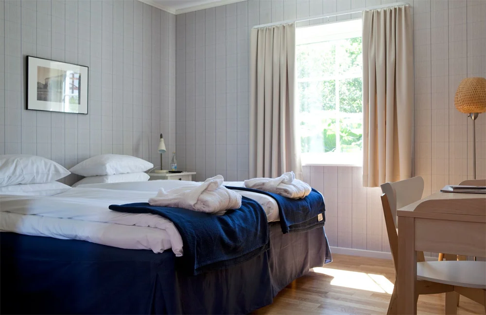 Une chambre d'hôtel soignée à Wallby Säteri comprend deux lits jumeaux avec des couvertures bleues, des peignoirs blancs, une fenêtre avec des rideaux beiges, un bureau et une chaise. L'espace accueillant est baigné de lumière naturelle.