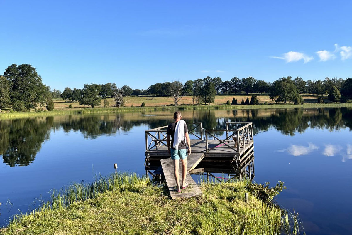 Une personne marche sur un quai en bois vers une petite plate-forme sur un lac calme à Wallby Säteri, entouré d'herbe verte, d'arbres et d'un ciel bleu clair.