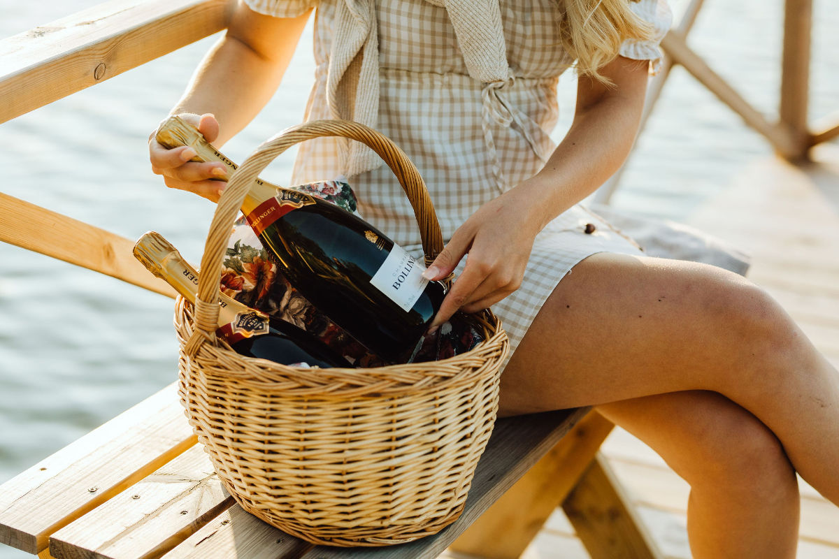Une personne vêtue d'une robe à carreaux est assise sur un banc en bois au bord de l'eau à Wallby Säteri, tenant un panier en osier rempli de bouteilles de vin mousseux.