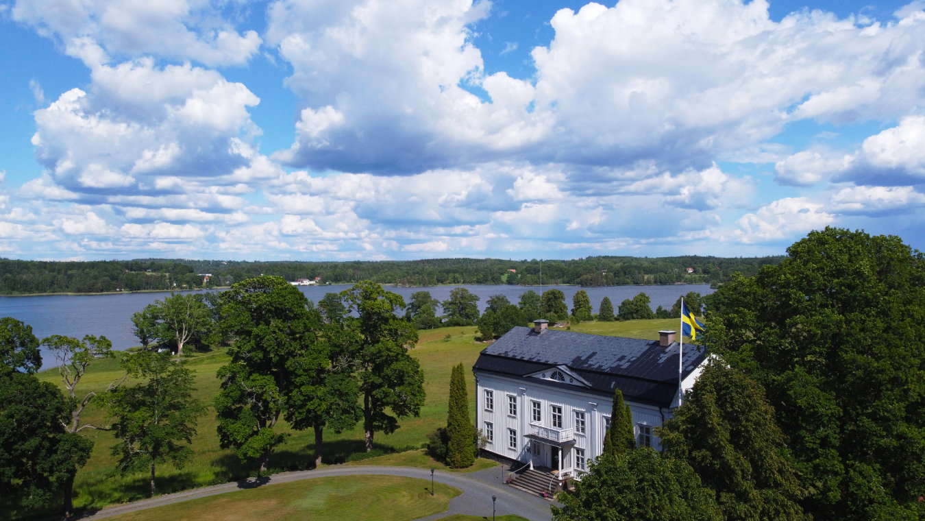Un grand bâtiment blanc, Wallby Säteri, avec un drapeau suédois, se trouve parmi les arbres près d'une rivière sous un ciel partiellement nuageux.