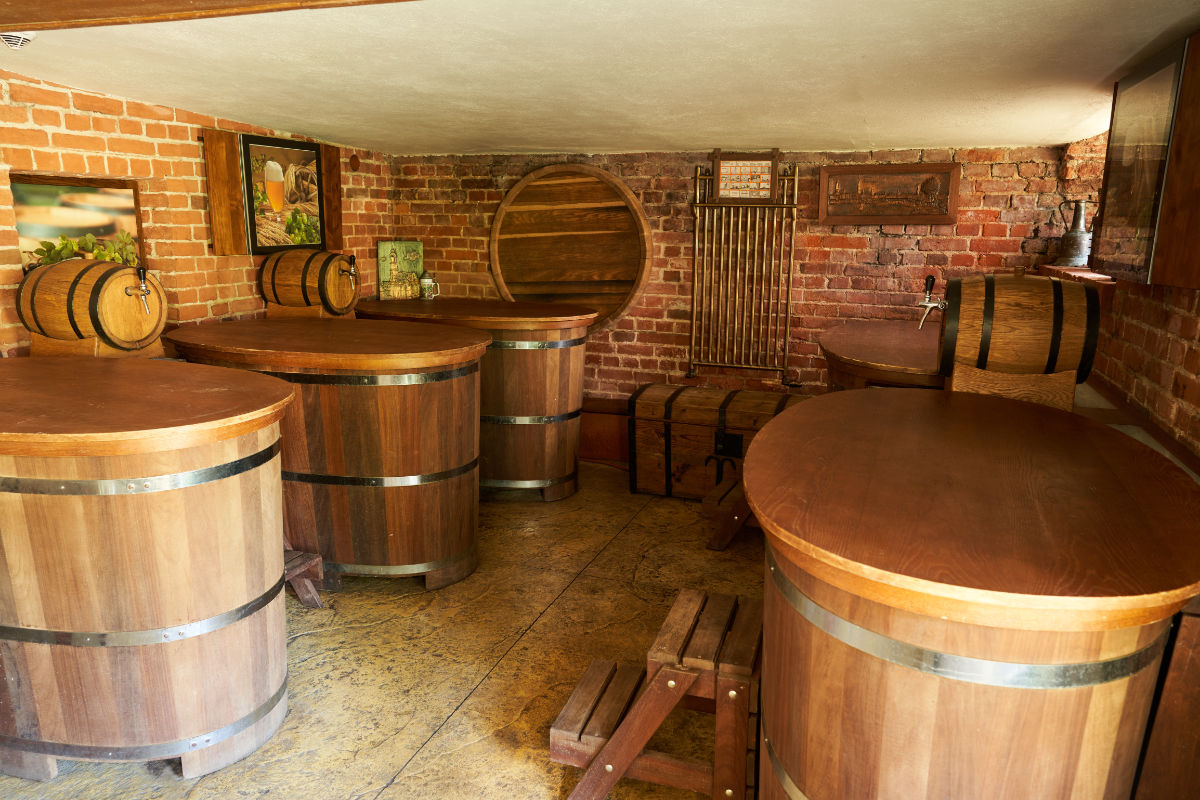 Ein Raum mit mehreren großen Holzfässern und Tischen, Backsteinwänden, Gemälden und Metallvorrichtungen, der an eine traditionelle Brauerei oder Brennerei erinnert.
