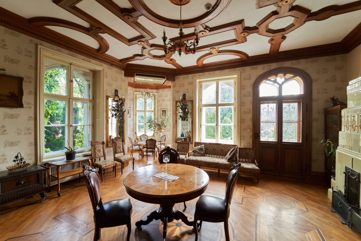 Elegantes, lichtdurchflutetes Wohnzimmer im Vintage-Stil mit verschnörkelter Holzdecke, großen Fenstern, antiken Möbeln und Parkettboden.