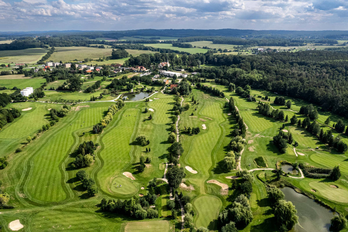Luftaufnahme eines großen Golfplatzes mit grünen Fairways, Sandbunkern, Wasserspielen, Bäumen und Gebäuden in der Nähe, umgeben von Feldern und Wald.