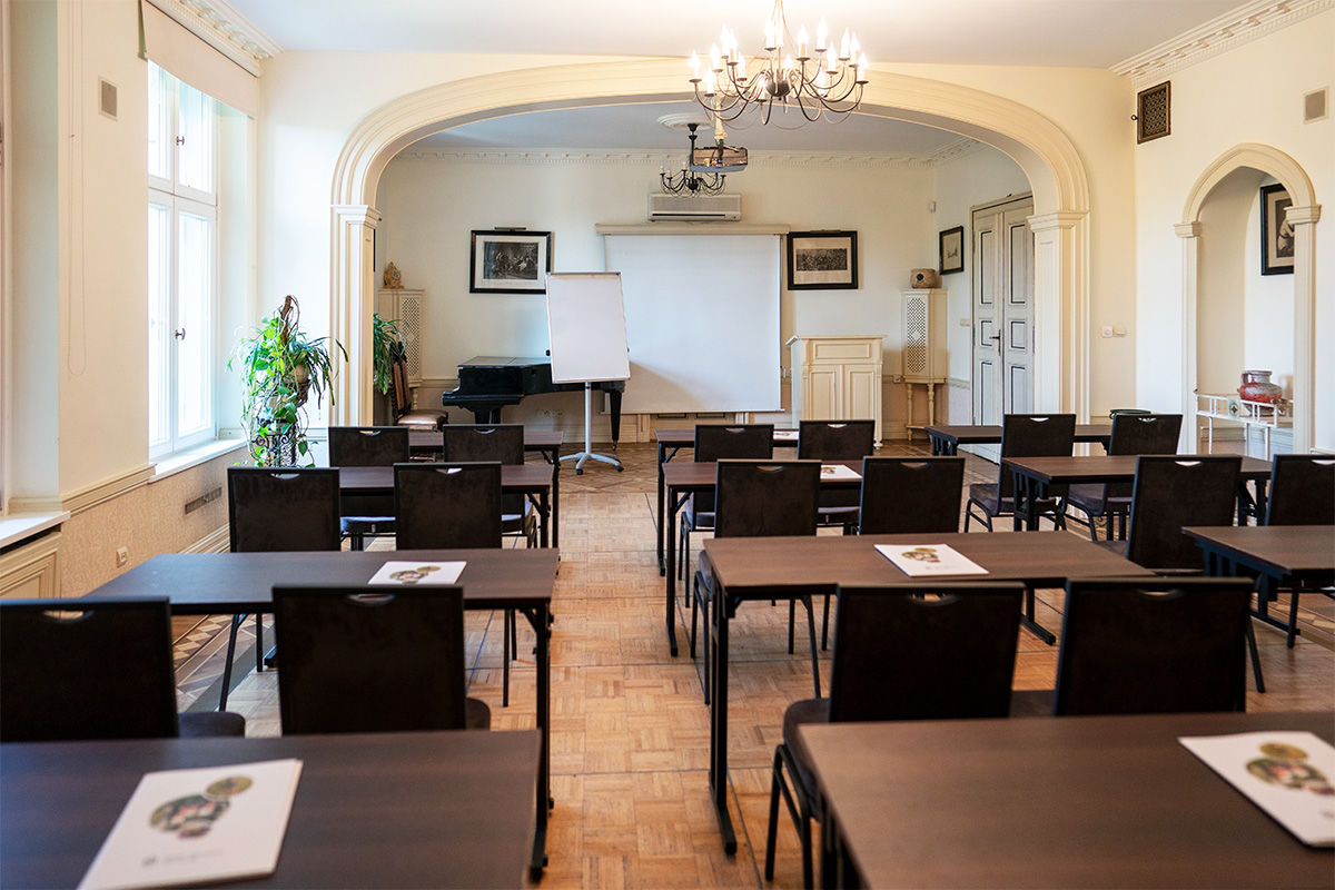 Ein Klassenzimmer mit Reihen von Tischen und Stühlen, einem Flipchart, einem Klavier und Dokumenten auf jedem Tisch. Der Raum ist durch große Fenster und einen Kronleuchter gut beleuchtet.
