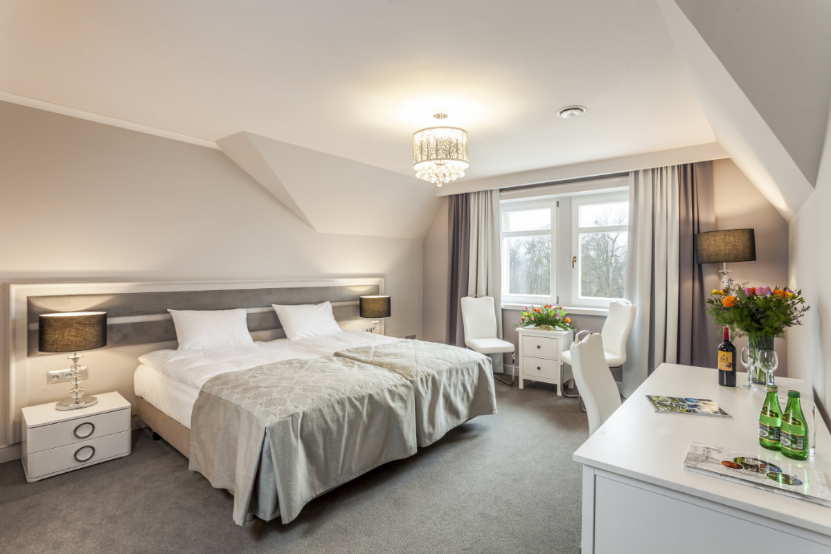 Ein Hotelzimmer mit einem Doppelbett, zwei Nachttischen mit Lampen, einem Schreibtisch mit Blumen und Getränken, zwei Stühlen am Fenster und neutraler Einrichtung.