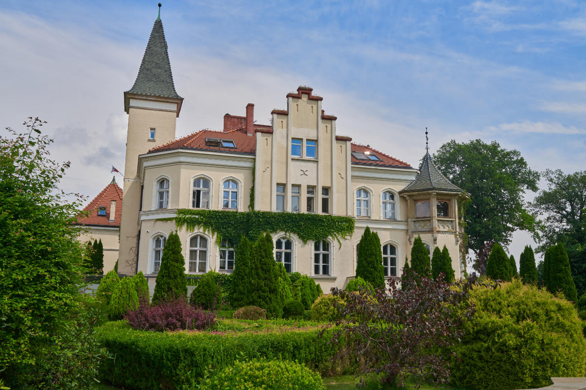 Grande edificio storico con torretta, edera sulla facciata e giardino antistante, sotto un cielo parzialmente nuvoloso.