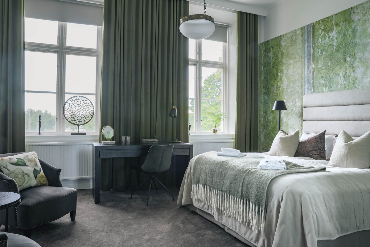 A modern bedroom inspired by Gysinge Manor features green curtains, beige and green bedding, a wall mural, a dark desk with a chair, and large windows that fill the space with natural light.