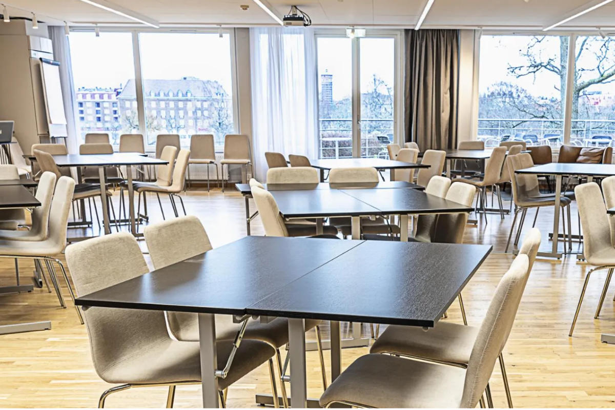 A modern, empty cafeteria with wooden floors, light-colored chairs, and dark tables, featuring large windows and an urban view—perfect for gatherings at Albert Hotel or business stays at albert kök hotell & konferens in Sweden.