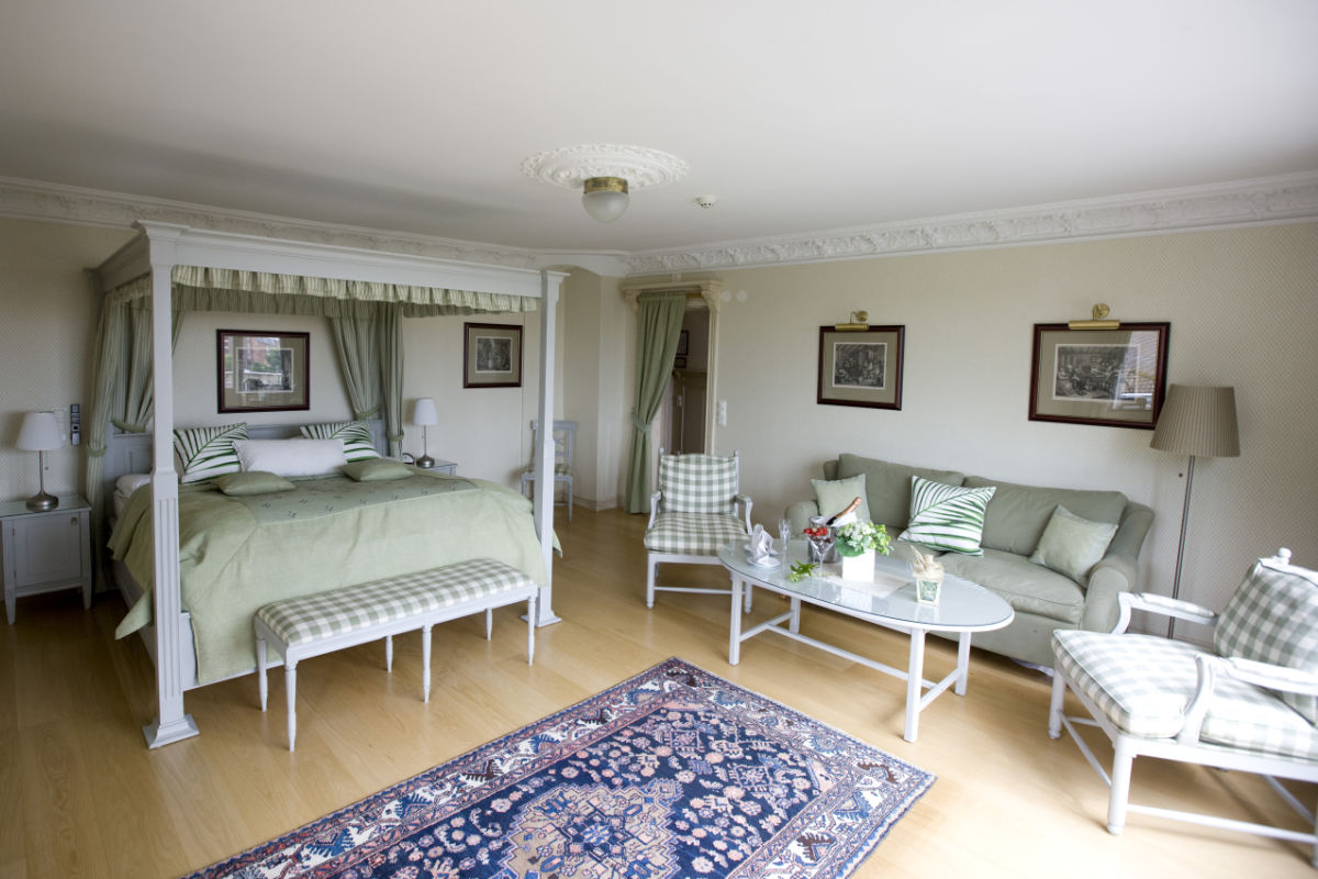 A spacious, well-lit bedroom at Albert Hotel features a canopy bed, green and white furniture, a sofa, armchairs, coffee table with flowers, and framed art on cream walls. A cozy rug rests on the wooden floor—a perfect stay in hotel Sweden.