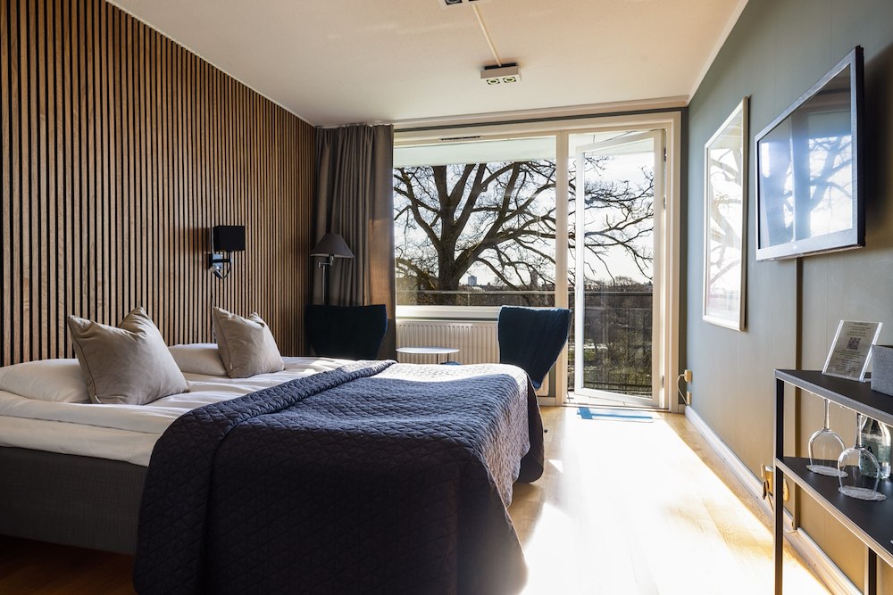 Modern hotel room at Albert Hotel featuring a double bed, wooden accent wall, wall-mounted TV, large window, and a balcony overlooking trees—perfect for a relaxing stay at this renowned hotel in Sweden.