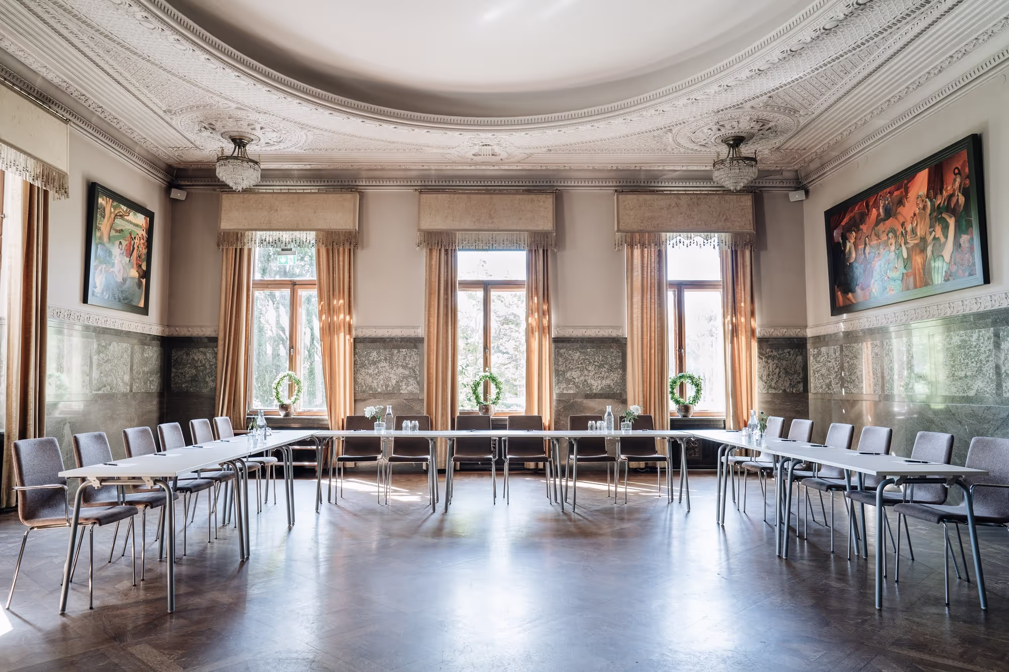 Una gran sala de reuniones ornamentada en el castillo de Bjertorp, con techos altos, mesas dispuestas, sillas vacías y obras de arte en las paredes, iluminada por la luz natural de altas ventanas.