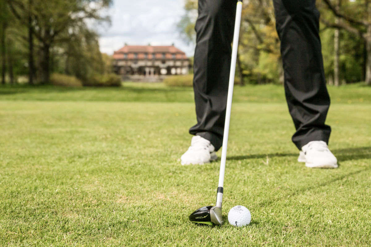 Una persona con pantalones negros y zapatos blancos se encuentra en un campo de golf cerca del castillo de Bjertorp, preparándose para golpear una pelota de golf con un palo; al fondo se ve el edificio del castillo.