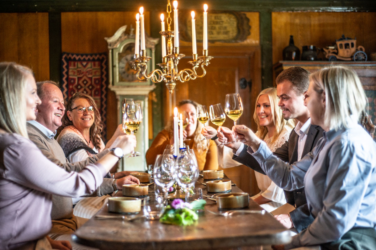 Acht Personen sitzen um einen Esstisch in Brösarps Gästgifveri & SPA und stoßen bei warmem Kerzenlicht und köstlichem Essen mit ihren Weingläsern an.