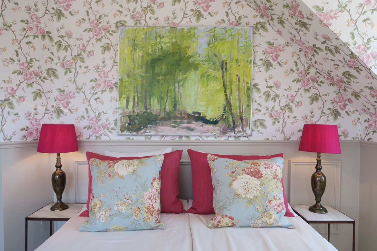 Ein Bett mit geblümten Kissen, flankiert von zwei Lampen mit rosafarbenen Schirmen, steht unter einem Gemälde eines grünen Waldes an einer Wand mit Blumentapete in Brösarps Gästgifveri & SPA.