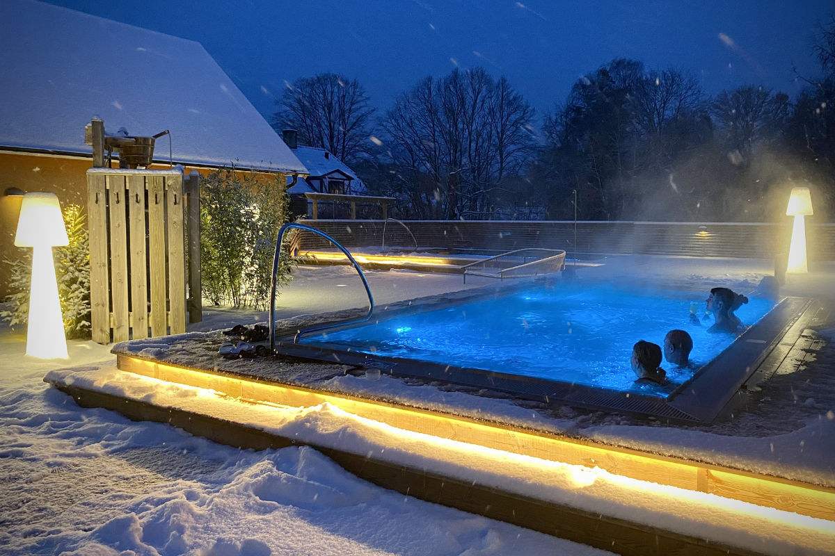 Beheiztes Freibad in Brösarps Gästgifveri & SPA, mit Menschen, die nachts schwimmen, umgeben von Schnee und beleuchtet von warmen Lichtern; im Hintergrund sind Bäume und Gebäude zu sehen.