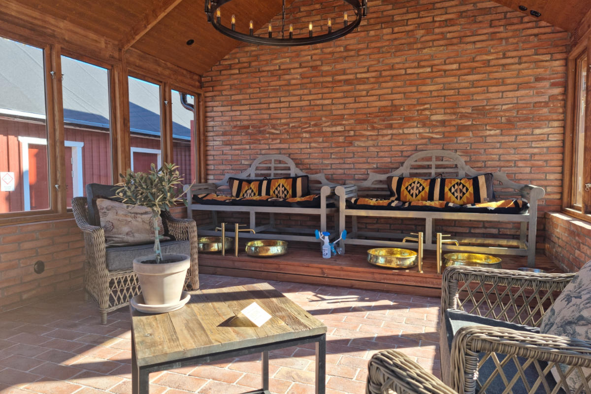 Ein sonnendurchfluteter Raum mit Backsteinwänden in Brösarps Gästgifveri & SPA verfügt über zwei Holzbänke mit Kissen, zwei Metallbecken, Korbstühle und einen kleinen Tisch mit einer Topfpflanze.