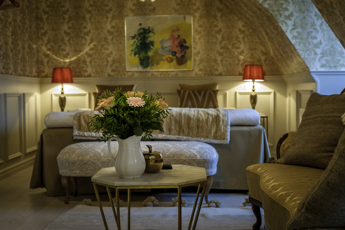 Ein gemütliches, warm beleuchtetes Schlafzimmer im Brösarps Gästgifveri & SPA verfügt über gemusterte Tapeten, ein Doppelbett, rote Lampen, einen kleinen Tisch mit einer weißen Blumenvase und einen goldenen Sessel.