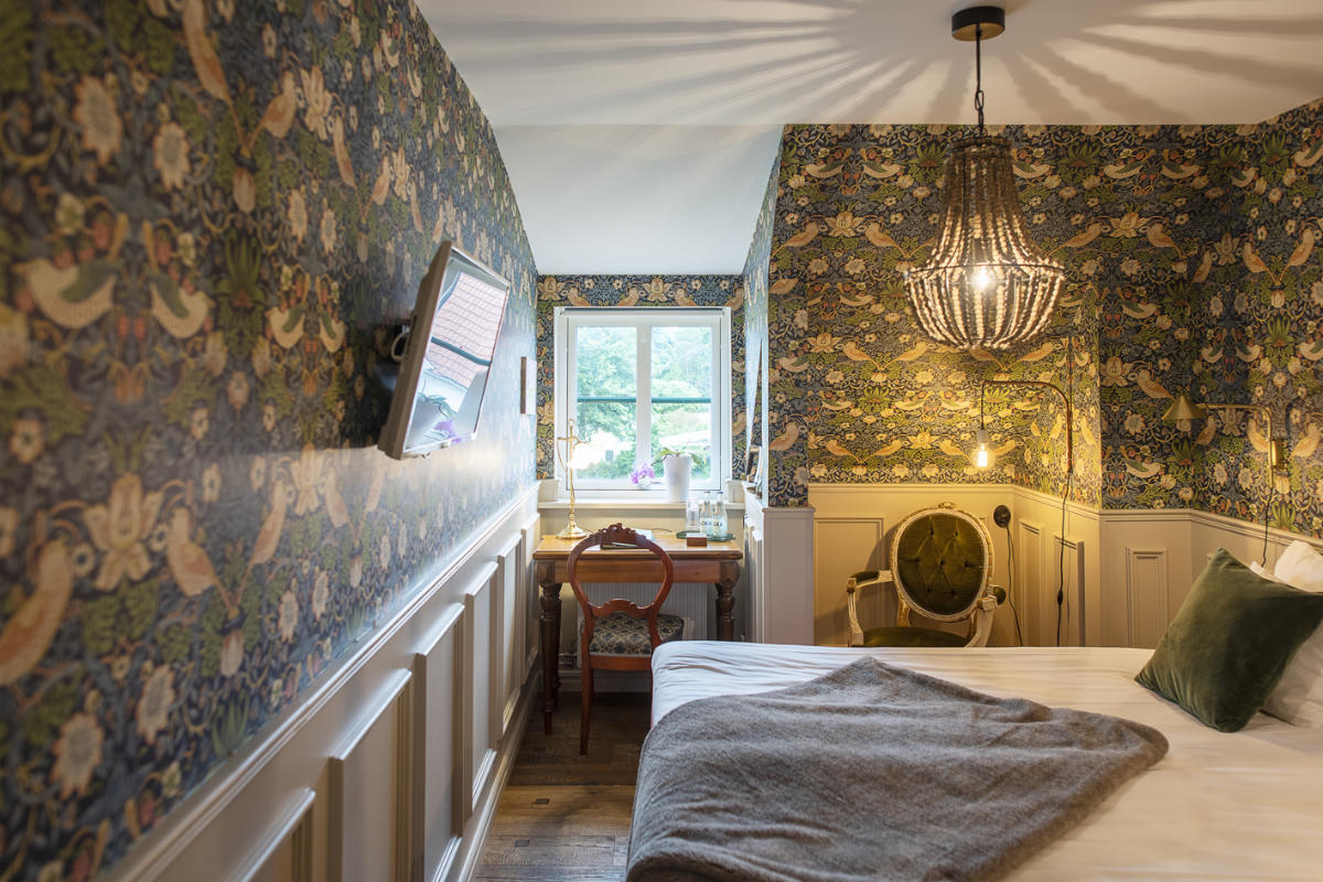 Ein gemütliches Schlafzimmer im Brösarps Gästgifveri & SPA mit Blumentapete, einem Bett mit weißer Bettwäsche, einem grünen Kissen, einer grauen Decke, einem an der Wand montierten Fernseher, einem Kronleuchter und einem Schreibtisch am Fenster, in das Tageslicht fällt.