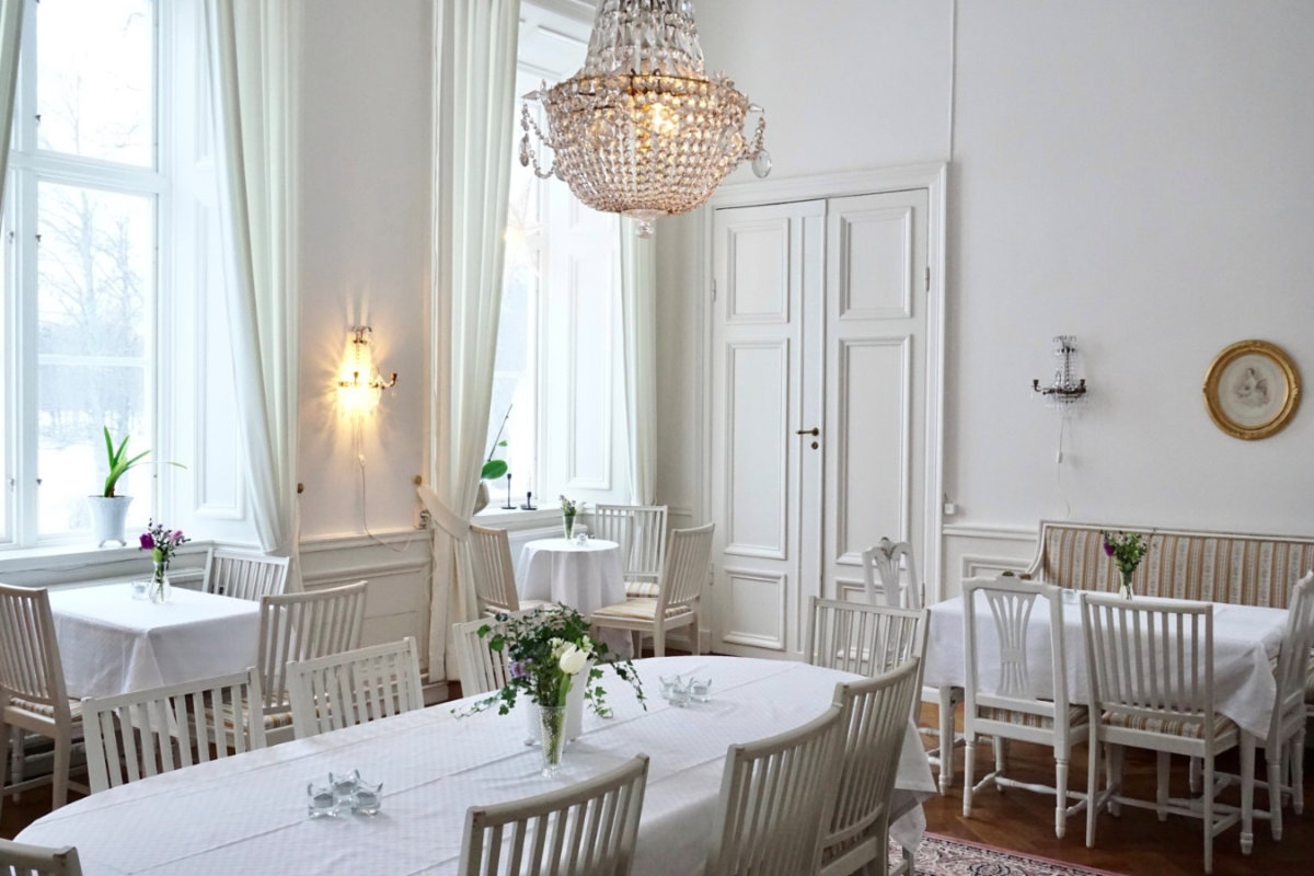 Bright dining room at Gysinge Manor with white walls, white tables and chairs, a crystal chandelier, large windows, and minimal floral decorations on the tables.
