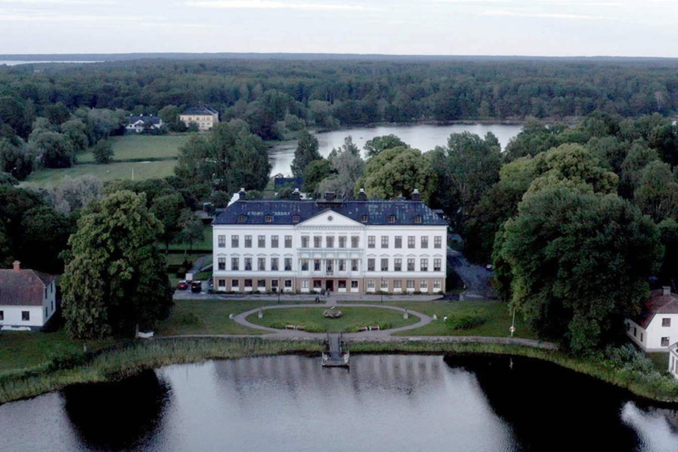 Gysinge Manor, a large white building with a symmetrical facade, stands by a lake surrounded by trees and smaller structures, with a forested area in the background.