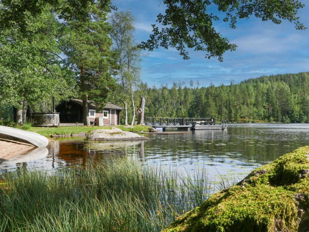 A lakeside cabin with a dock sits among trees beside calm water, under a blue sky. Near Hennickehammars Herrgård, a canoe rests on the shore, surrounded by forest and greenery.