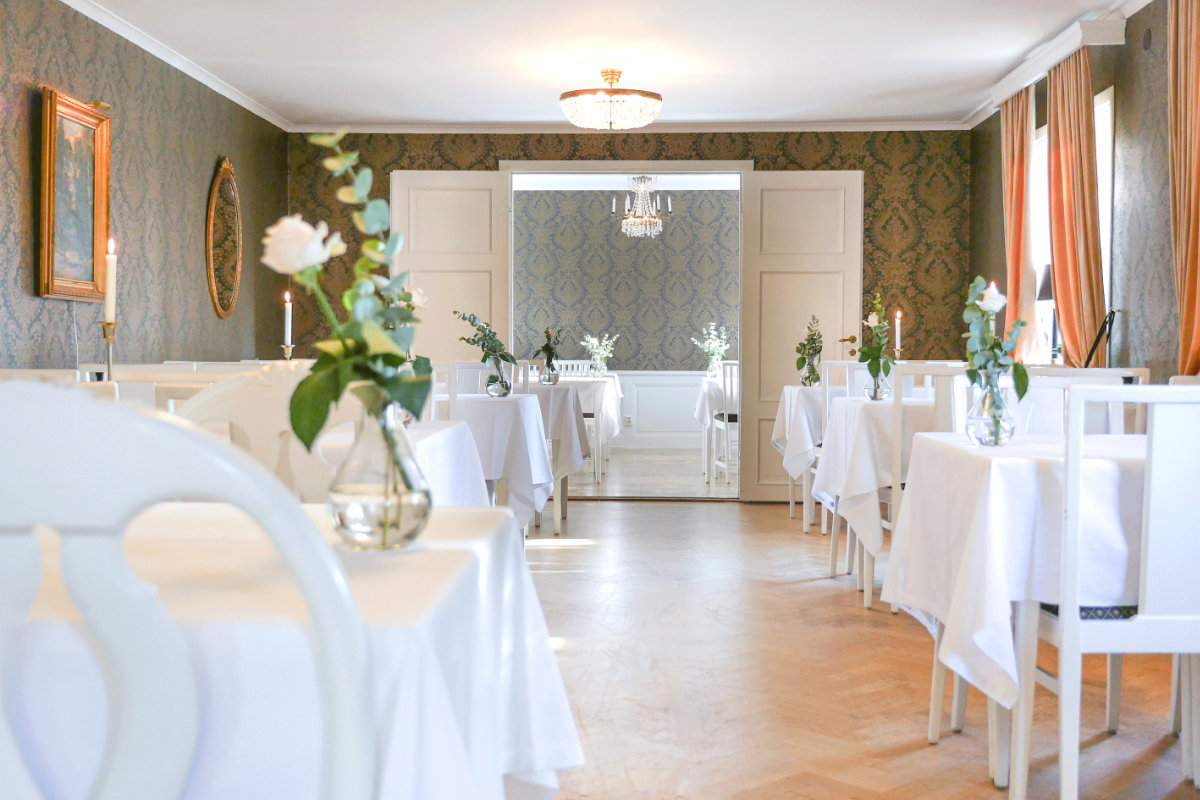 Elegant restaurant dining room at Hennickehammars Herrgård with white tablecloths, floral centerpieces, wooden floor, patterned wallpaper, and chandeliers, arranged for a formal seating.