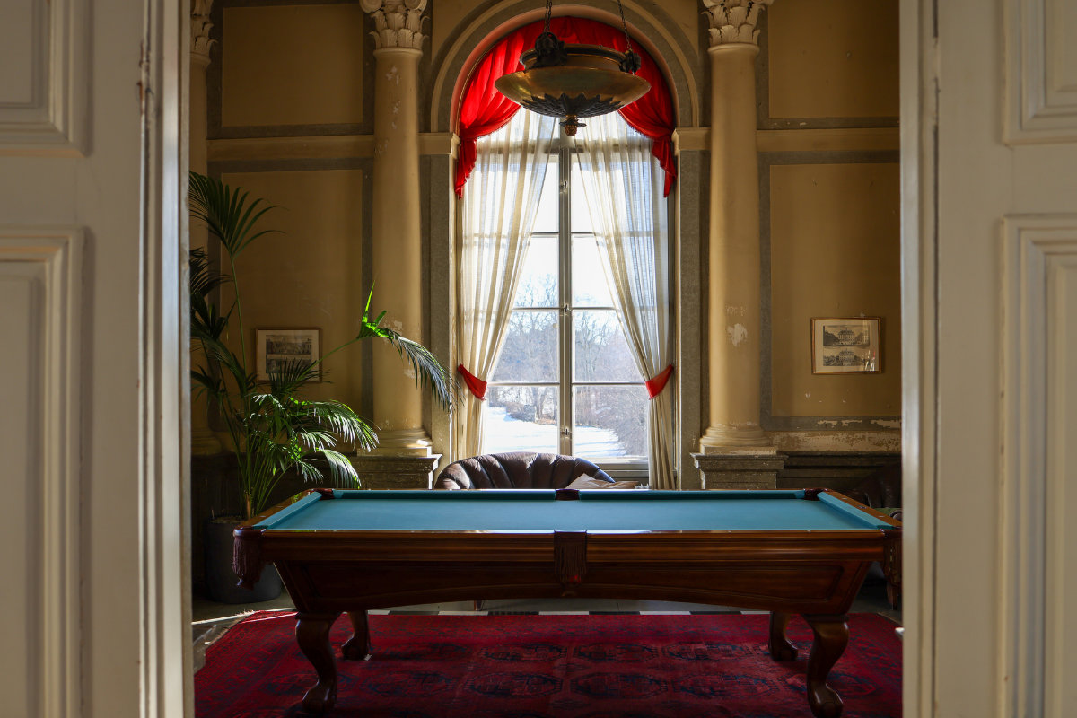 Un tavolo da biliardo poggia su un tappeto rosso in una stanza con alte finestre, tende rosse, colonne, una pianta e quadri incorniciati alle pareti.