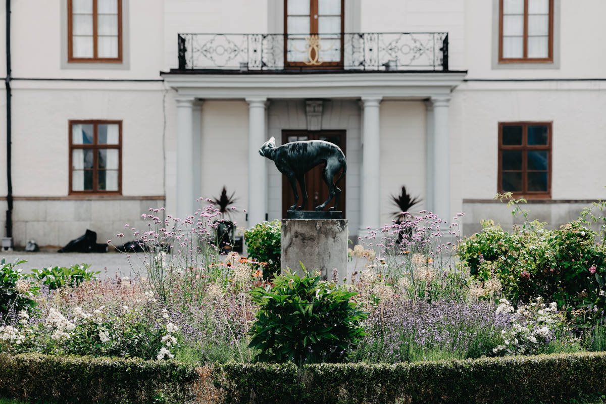 Una statua di animale in bronzo si trova su un piedistallo al centro di un giardino fiorito di fronte a un grande edificio con pareti bianche e finestre con cornice marrone.