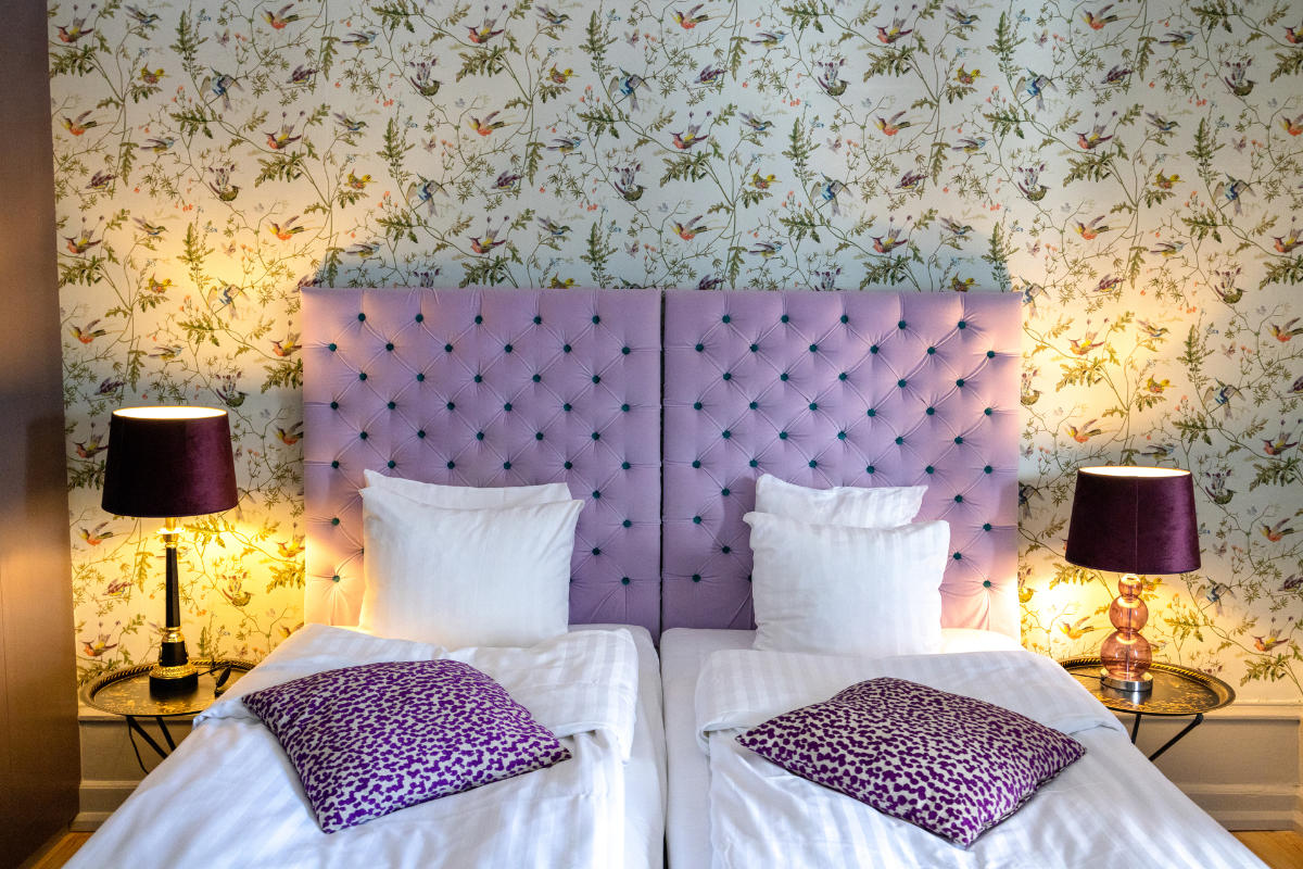 Due letti singoli con lenzuola bianche e cuscini a fantasia viola, una testata lilla con inserti, due comodini e lampade su carta da parati floreale.