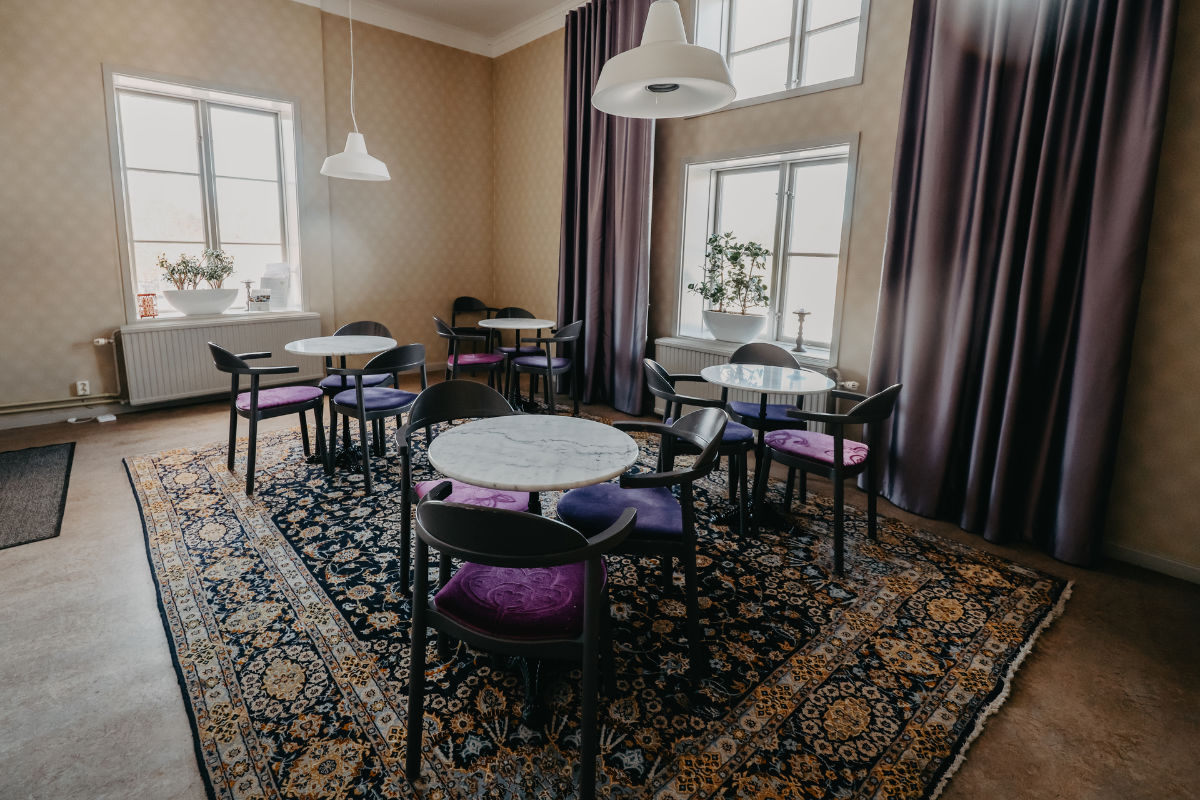 Una piccola area caffè con tavoli rotondi in marmo, sedie nere con cuscini viola e grandi finestre con tende viola, il tutto su un tappeto a fantasia.