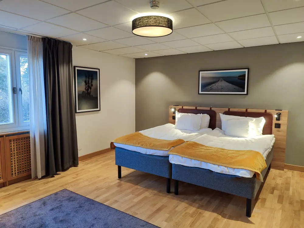 Una habitación de hotel en Skåvsjöholm Hotell & Konferens con dos camas individuales juntas, cada una con ropa de cama blanca y una manta amarilla, suelos de madera clara, una ventana con cortinas y fotos enmarcadas en las paredes.