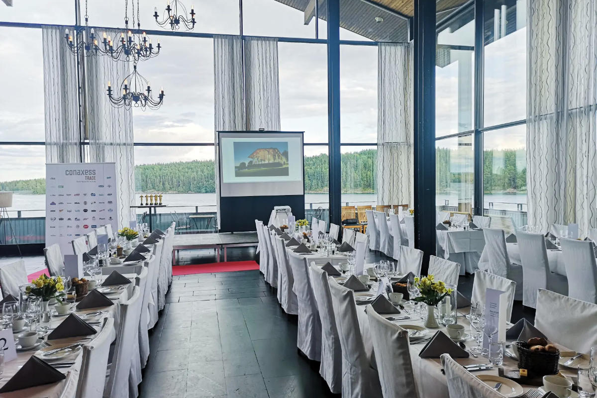 El comedor formal del Skåvsjöholm Hotell & Konferens cuenta con dos mesas largas, sillas blancas, centros de mesa florales y una pantalla de proyección en una sala acristalada con vistas al lago y al bosque.