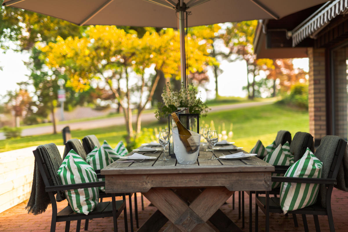 Ein hölzerner Esstisch im Freien des Skåvsjöholm Hotell & Konferens ist mit Tellern, Gläsern und grün gestreiften Kissen auf Stühlen unter einem großen Sonnenschirm gedeckt, umgeben von üppigem Grün und Bäumen.