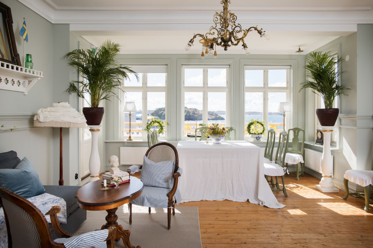 Un salon lumineux avec du parquet, des meubles classiques, des plantes d'intérieur et de grandes fenêtres donnant sur une vue pittoresque sur l'extérieur, rappelant l'élégance douillette de l'hôtel Strandflickorna. Une table blanche et des chaises sont disposées près des fenêtres.