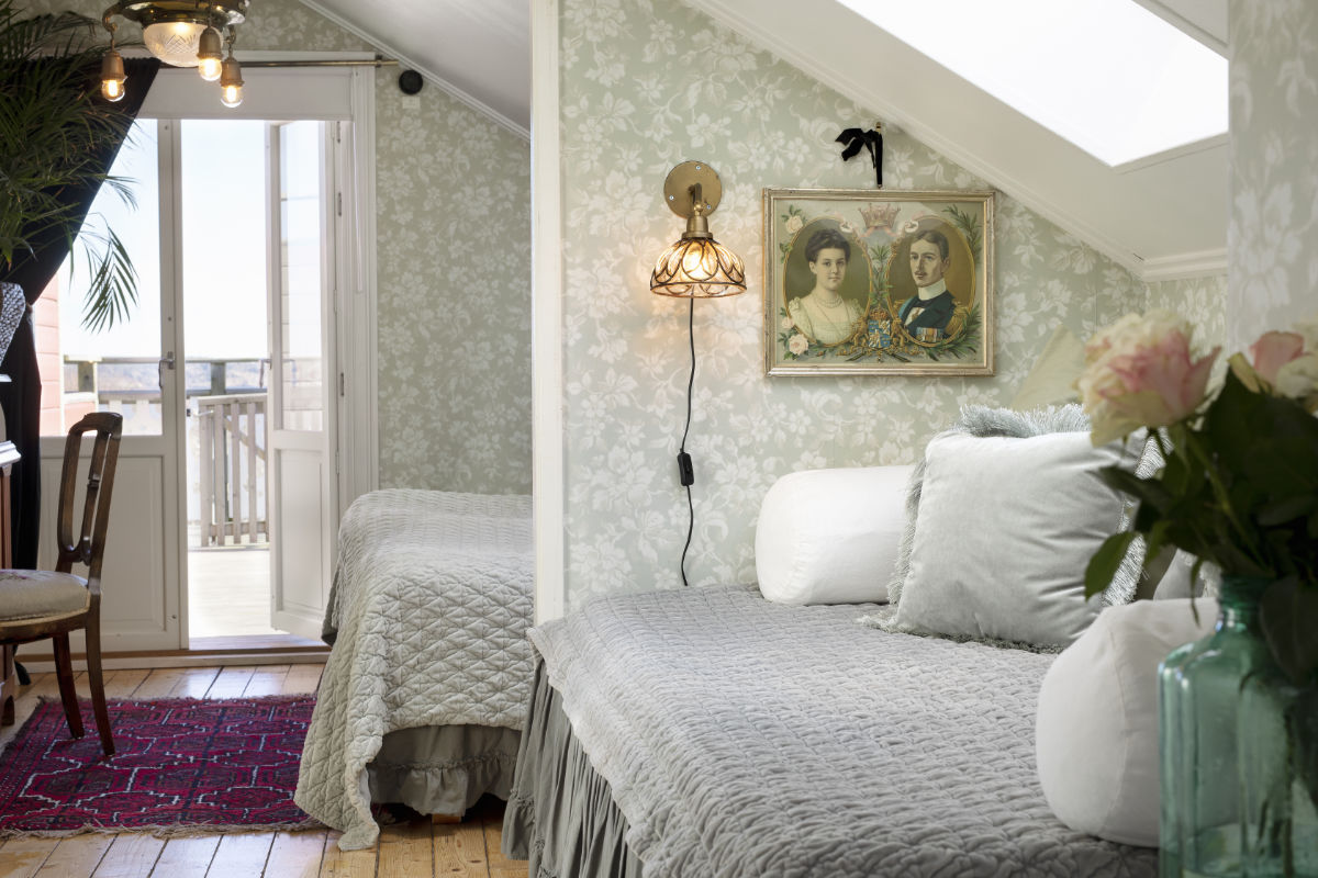 La chambre mansardée de l'hôtel Strandflickorna présente un papier peint floral, un lit de repos avec une literie grise et blanche, un portrait encadré, des fleurs sur une table et une porte menant à un balcon.