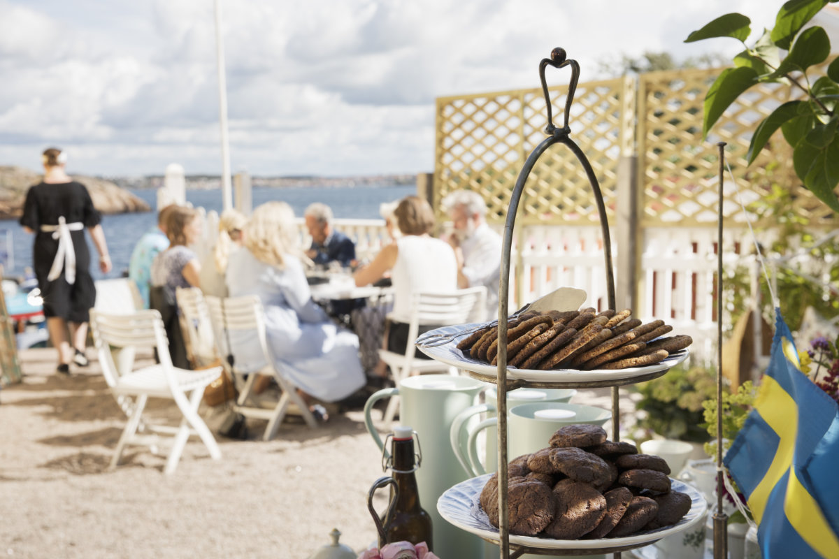 Une assiette de biscuits sur un support en gradins au premier plan à l'hôtel Strandflickorna, avec des personnes assises à des tables et des chaises blanches en plein air au bord de l'eau, sous un ciel nuageux.