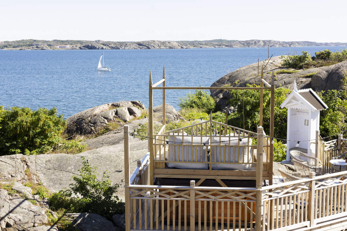 À l'hôtel Strandflickorna, une terrasse en bois avec des sièges surplombe un terrain rocheux et une mer bleue calme, avec un voilier au loin et des îles côtières à l'horizon.