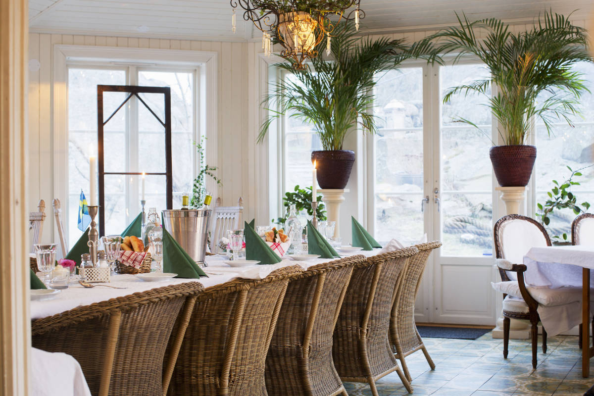 Une longue table à manger avec des chaises en osier est garnie de verres, d'assiettes, de serviettes vertes et de fleurs dans une pièce lumineuse de l'hôtel Strandflickorna, qui dispose de grandes fenêtres, de plantes en pot et d'une lumière naturelle abondante.