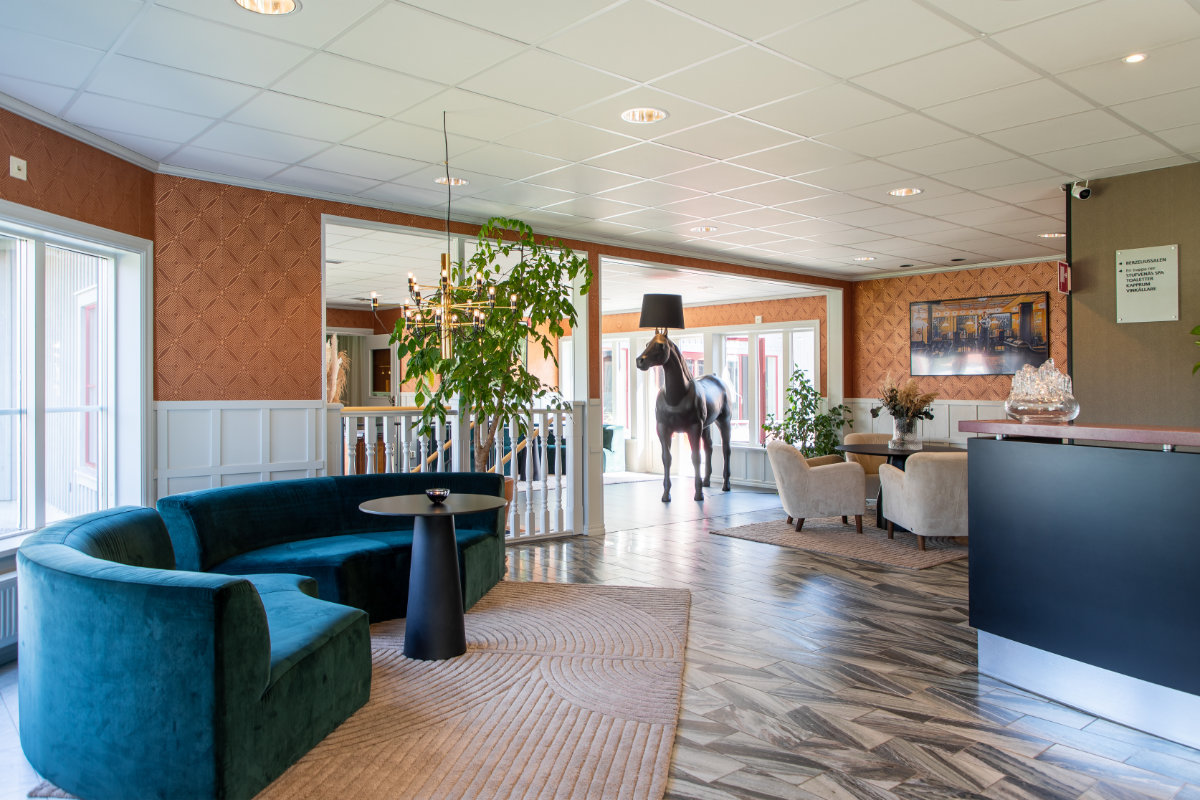 Moderno vestíbulo de hotel en Stufvenäs Gästgifveri con un sofá de terciopelo verde, mesa redonda, sillones, exuberantes plantas de interior y una lámpara en forma de caballo cerca de grandes ventanales. El mostrador de recepción se ve a la derecha.