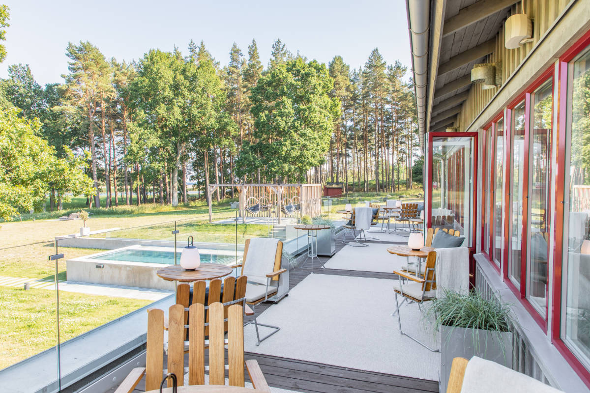 Zona de estar al aire libre con sillas y mesas de madera en una terraza de Stufvenäs Gästgifveri, con vistas a un patio de césped con piscina, árboles y un cielo despejado al fondo.
