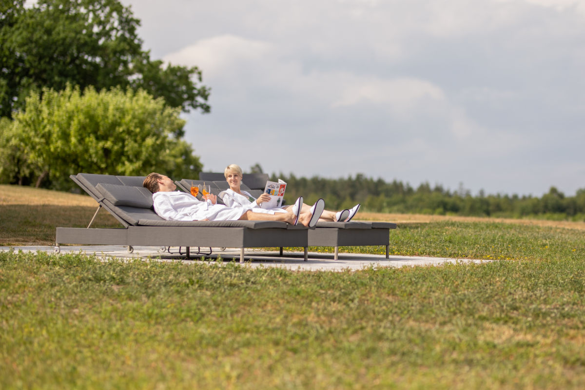 Dos personas vestidas con batas blancas se relajan en tumbonas al aire libre en Stufvenäs Gästgifveri; una lee una revista mientras la otra sostiene una bebida. Disfrutan de un tranquilo patio rodeado de césped y árboles bajo un cielo nublado.