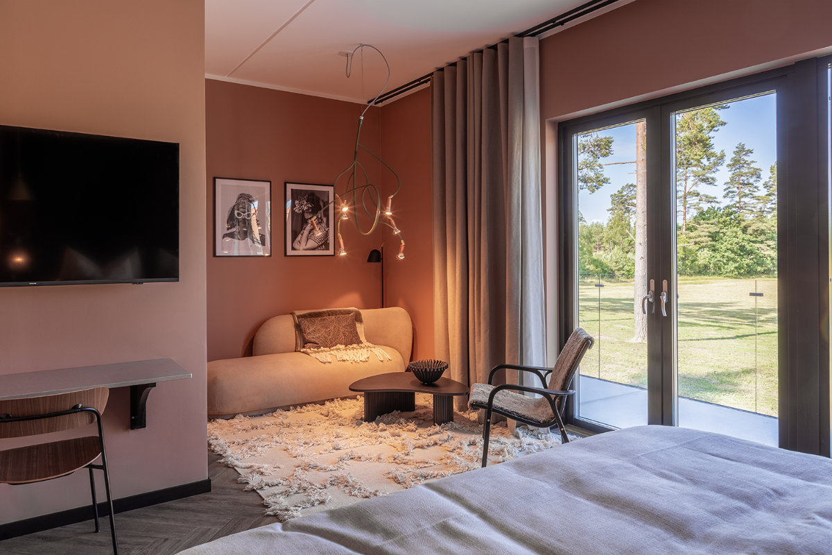 Moderna habitación de hotel en Stufvenäs Gästgifveri con sofá beige, obras de arte en la pared, televisor y puertas de cristal que dan a una zona exterior cubierta de hierba. Los tonos neutros y la iluminación tenue crean un ambiente acogedor.