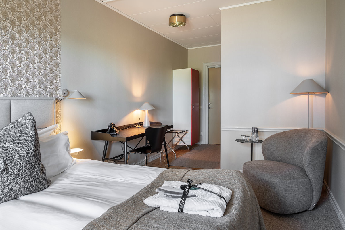 La habitación de hotel del Stufvenäs Gästgifveri está perfectamente equipada con cama doble, escritorio con silla, tumbona y armario. Las toallas están dobladas sobre la cama y las lámparas proporcionan una iluminación suave.