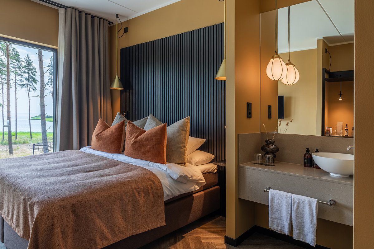 Una moderna habitación en Stufvenäs Gästgifveri cuenta con una cama doble con varias almohadas, ropa de cama de color topo, lámparas colgantes y un tocador con lavabo y toalla, junto a una ventana con vistas a los árboles y al agua.