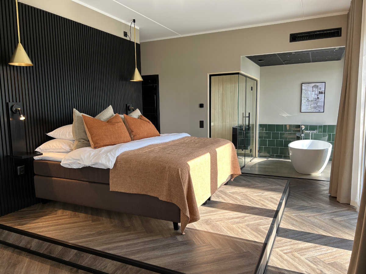 Moderna habitación de hotel en Stufvenäs Gästgifveri con cama de matrimonio, ropa de cama naranja, lámparas colgantes, suelos de madera y cuarto de baño abierto con bañera exenta y azulejos verdes.