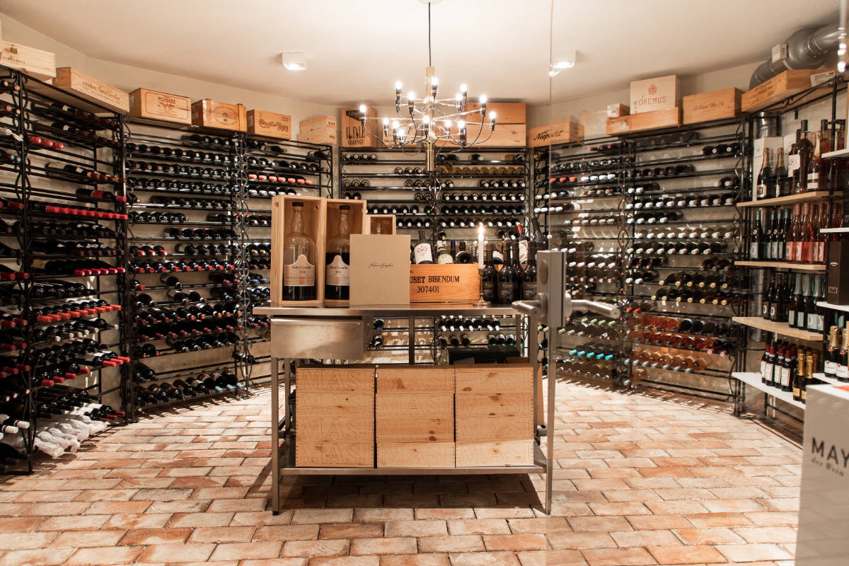 Una bodega en Stufvenäs Gästgifveri con estanterías llenas de botellas de vino, cajas de madera sobre una mesa central y una lámpara de araña en lo alto, todo ello sobre un rústico suelo de ladrillo.